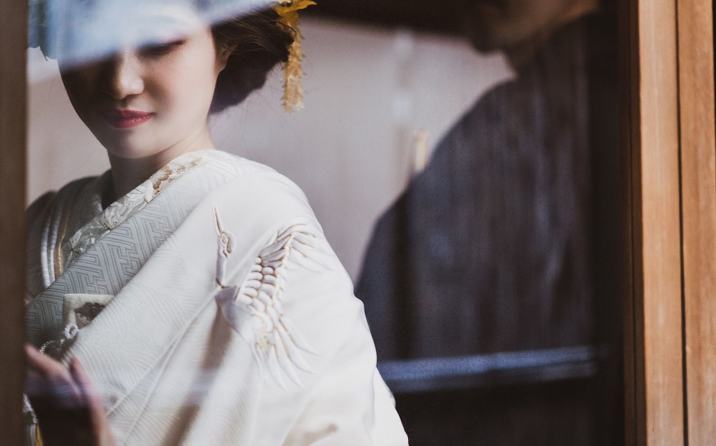 白い着物を着用し、目を閉じて穏やかに微笑む日本の花嫁のクローズアップポートレート。柔らかな光が顔を照らし、背景にはぼやけた人物のシルエットと木製の柱が見える