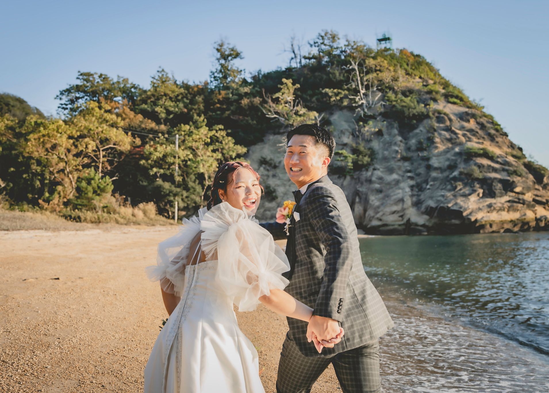 砂浜と海を背景に、白いウェディングドレスを着た花嫁とチェック柄のスーツを着た新郎が、緑豊かな木々と岩の崖の前で満面の笑みを浮かべている海岸線での結婚式の様子