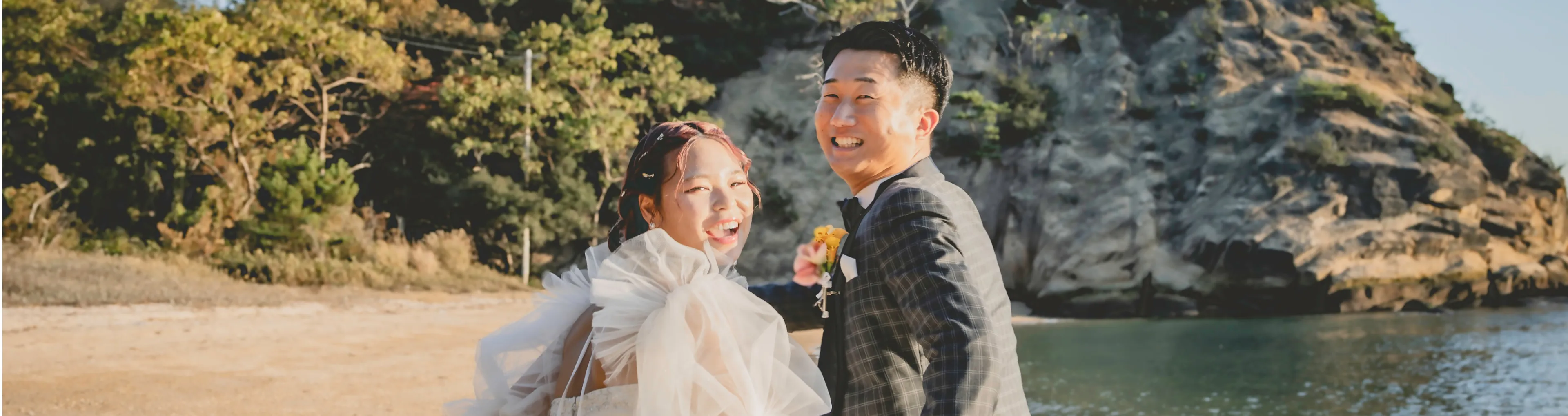 砂浜と海を背景に、白いウェディングドレスを着た花嫁とチェック柄のスーツを着た新郎が、緑豊かな木々と岩の崖の前で満面の笑みを浮かべている海岸線での結婚式の様子