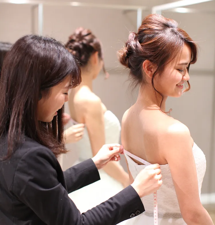 白いストラップレスのウェディングドレスを試着する花嫁と、肩のサイズを測っているスタッフの様子