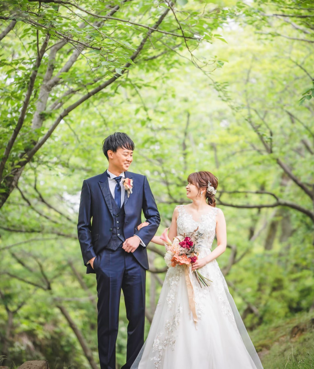 ブーケを手にした白いドレスの花嫁とダークスーツを着た花婿が、屋外の緑の木々の下に立ち、微笑みながら結婚式の日を見つめ合っている。.
