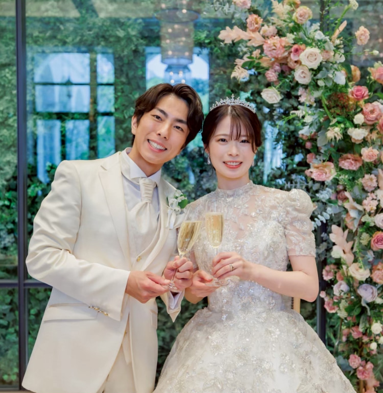 白いウェディングドレスを着た花嫁とクリーム色のタキシードを着た新郎が、シャンパンで乾杯している結婚式の様子。満面の笑みを浮かべ、背景にはピンクやクリーム色の花々で装飾されたアーチが見える