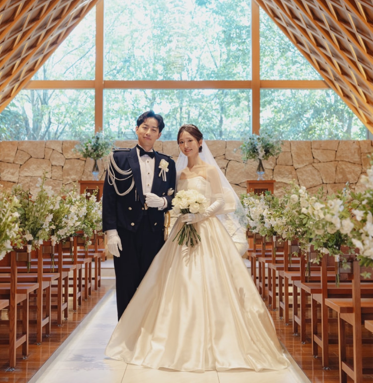 ダークスーツに白い手袋をはめた新郎の横に、白いガウンとベールをまとった新婦が立っている。花と木のアクセントで飾られた陽光降り注ぐチャペルで微笑むふたり。.
