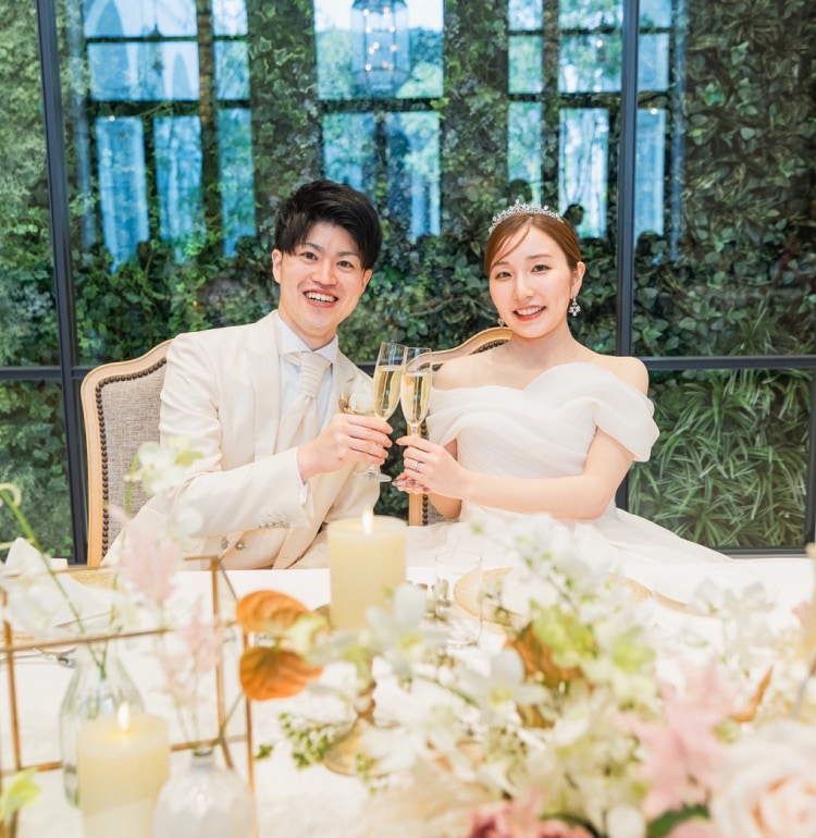 結婚式の衣装を着た新郎新婦が、飾られたテーブルに一緒に座り、笑顔でシャンパングラスを掲げて乾杯している。背景には大きな窓と豊かな緑。.