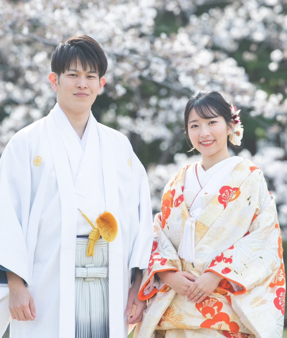 満開の桜を背景に、日本の伝統的な婚礼衣装に身を包んだ男女が並んで微笑んでいる。.