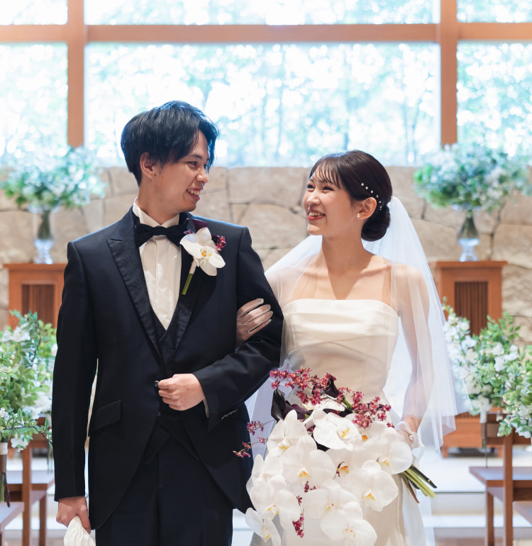 明るくエレガントな屋内で、花と緑に囲まれたバージンロードを腕を組んで歩きながら微笑み合う新郎新婦。花嫁は白い蘭のブーケを手にしている。.