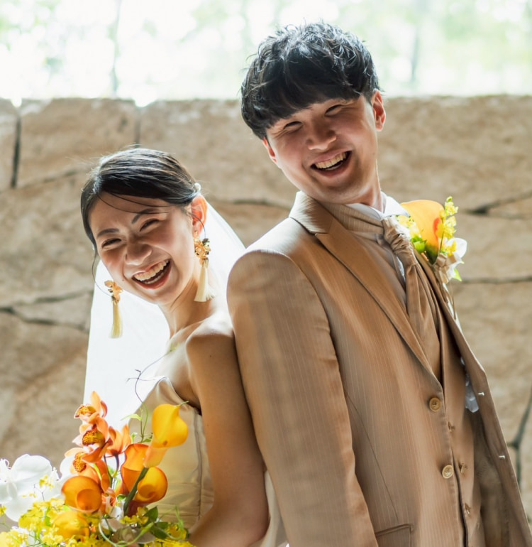 石壁と緑を背景に、ウエディングドレスに身を包み、黄色とオレンジの花束を手にした笑顔の新郎新婦が背中合わせに立っている。.