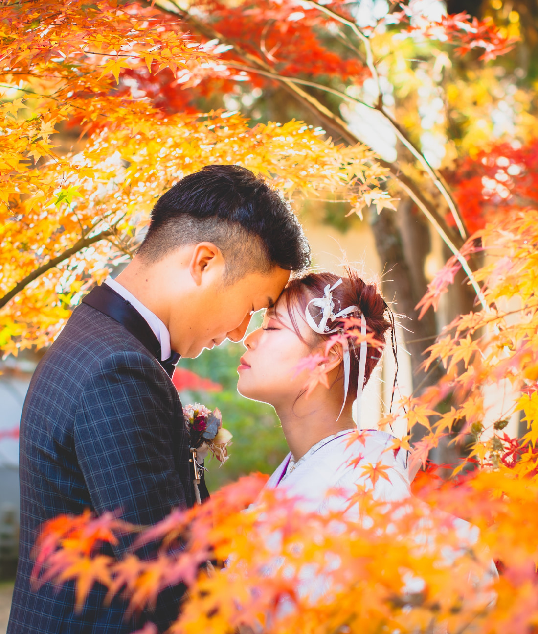赤、オレンジ、黄色など色とりどりの鮮やかな紅葉に囲まれ、結婚式の衣装を身にまとったカップルが仲良く立ち、愛おしそうに額を触れ合っている。柔らかな日差しが色とりどりの葉を透かしている。.