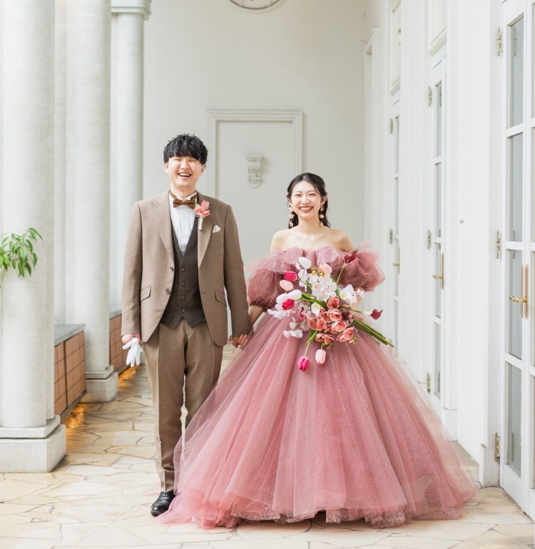 明るい廊下で手を取り合って微笑むカップル。新郎は日焼けしたスーツに蝶ネクタイ、新婦はピンクのボールガウンを着てピンクと白の花束を持っている。.