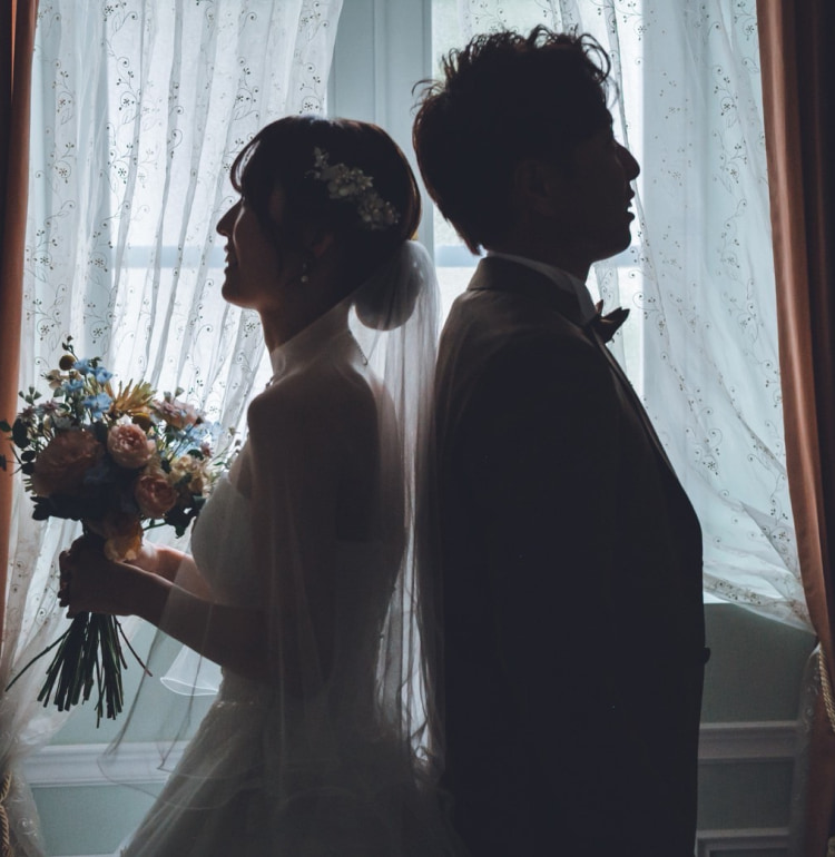 ブーケを持った花嫁と花婿が、シアーなカーテンの窓の前に背中合わせにシルエットで立っている。柔らかな自然光が花嫁のドレスとベールを際立たせている。.
