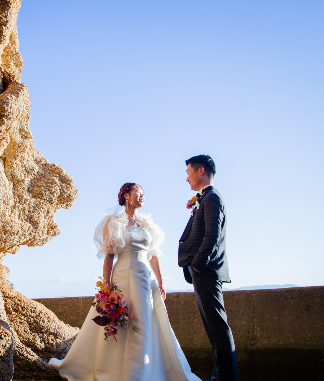 屋外の大きな岩のそばに立ち、微笑み合う新郎新婦。新婦は色とりどりの花束を持ち、新郎はスーツを着ている。背景の空は青く澄んでいる。.