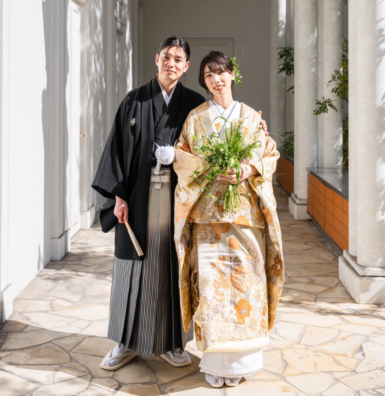 日本の伝統的な婚礼衣装に身を包んだカップルが微笑みながら立っている。新郎は黒い着物に袴、新婦は金と白の着物を着てブーケを持っている。二人は白い柱のある陽のあたる廊下でポーズをとっている。.
