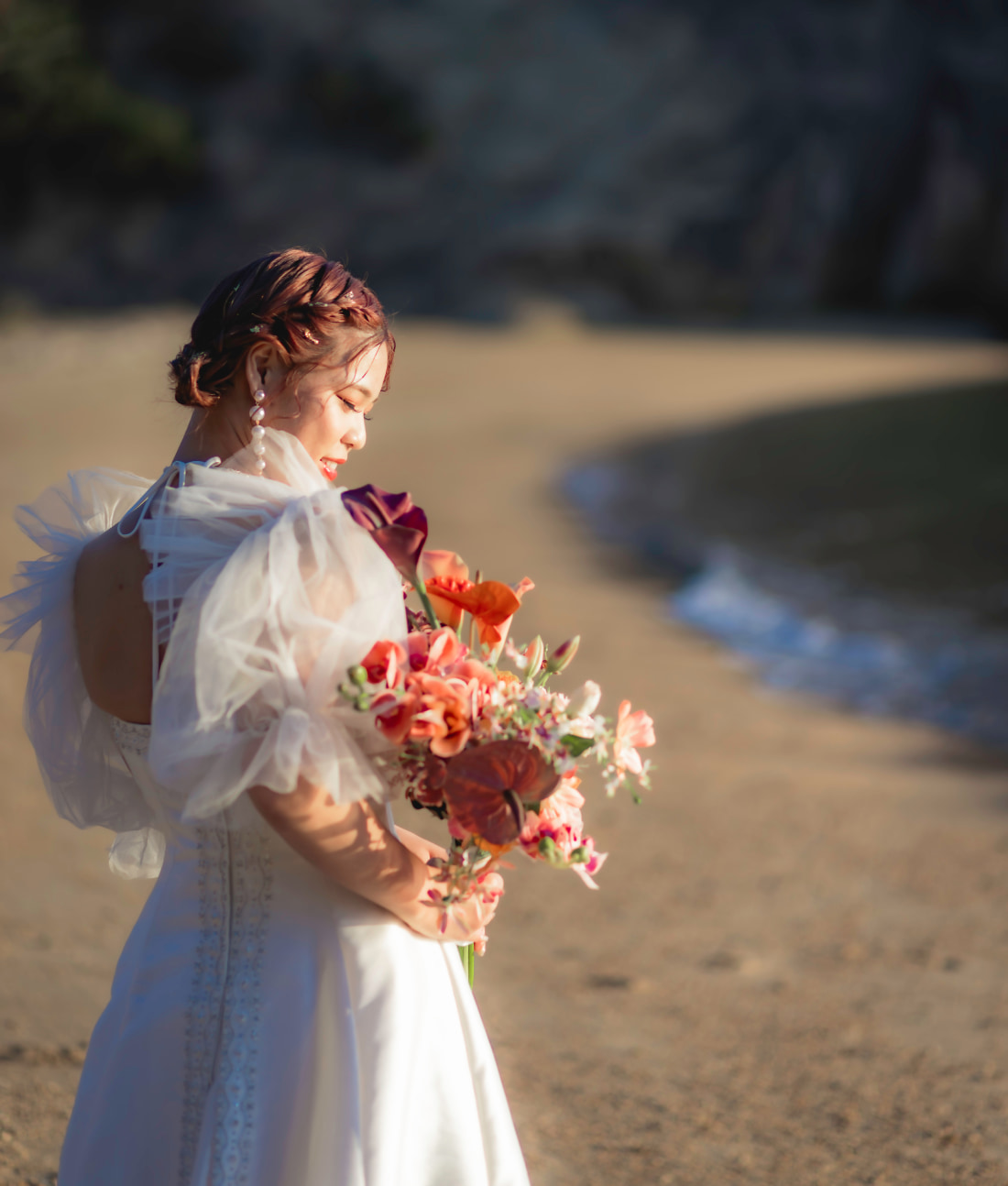 色とりどりのブーケを持った白いドレスの花嫁が砂浜に佇み、下を向いて優しく微笑んでいる。.