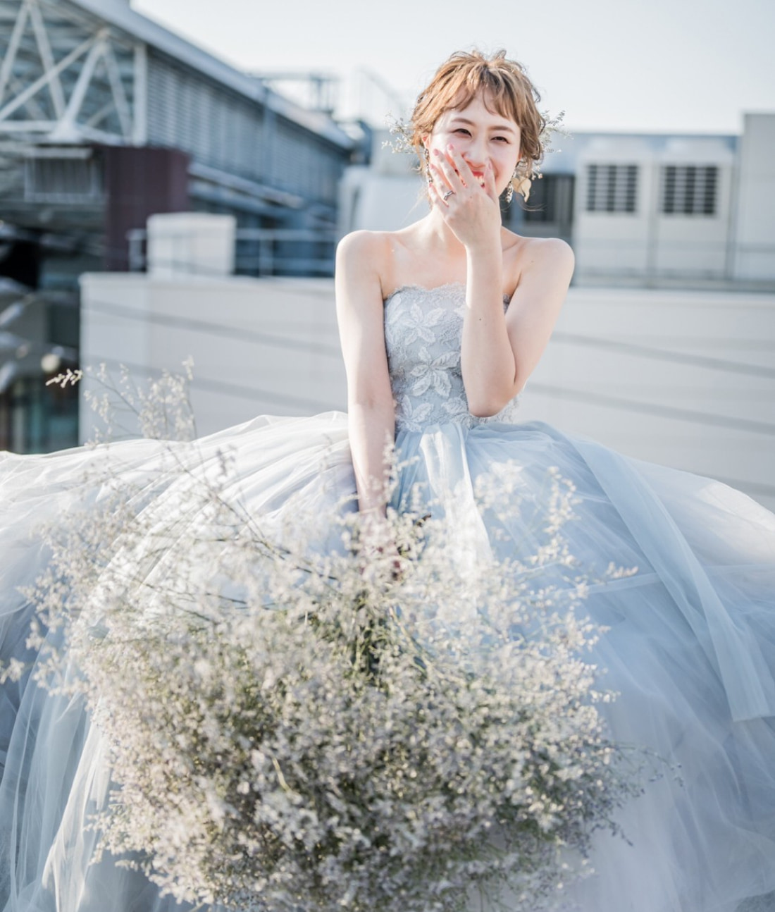 ストラップレスの水色のドレスに身を包んだ笑顔の花嫁が、繊細な白い花の大きなブーケを持ち、片手で口元を戯れに隠しながら屋外に立っている。背景には都会のビル群が見える。.