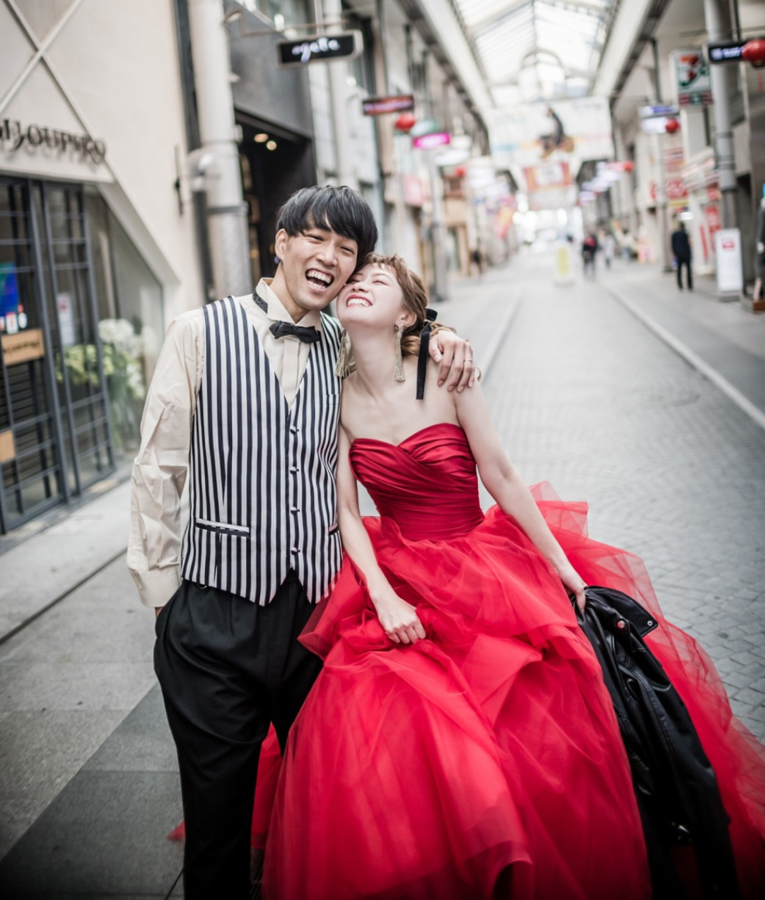 街角でポーズをとる楽しげなカップル。男性はストライプのベストに蝶ネクタイ、女性は鮮やかな赤のガウンを着て、黒いジャケットを持ちながら男性に寄りかかり、2人とも明るく笑っている。.