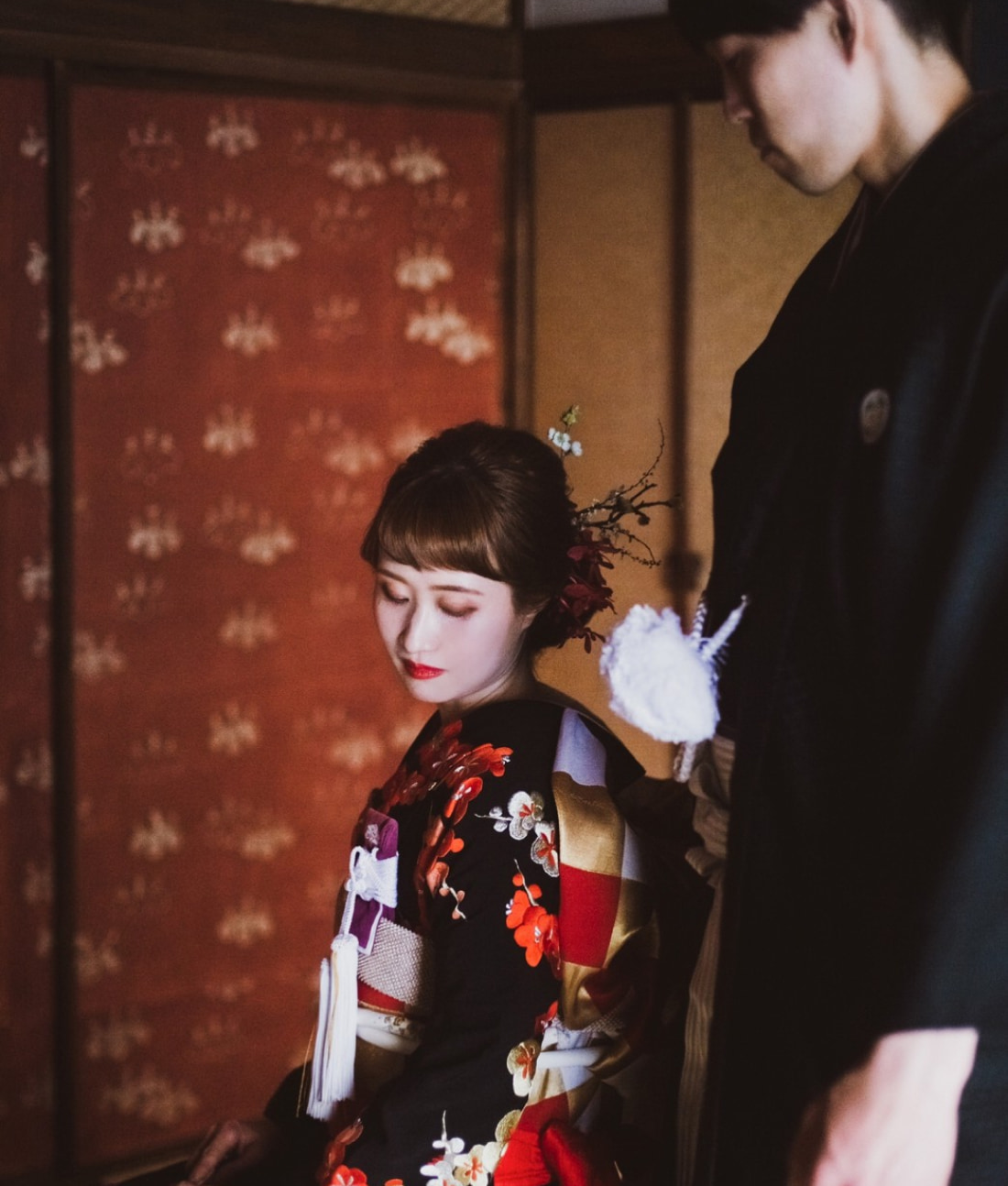 花柄の着物に髪飾りをつけた女性が下を向いて座り、その横に黒い伝統的な装いの男性が立っている。.