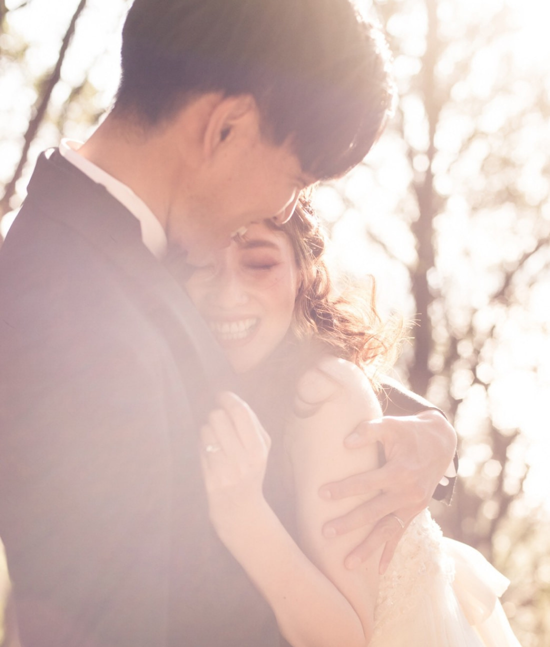 柔らかな日差しの中で抱き合い、楽しげに微笑むカップル。女性は白いドレス、男性はダークスーツという正装で、屋外の陽光に照らされた背景に囲まれている。.