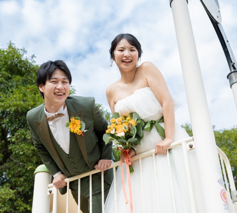 白いストラップレスのウェディングドレスを着た花嫁とオリーブグリーンのスリーピーススーツを着た新郎が、屋外の遊び場のような場所で、白い手すりにもたれかかりながらカメラに向かって満面の笑みを浮かべている様子