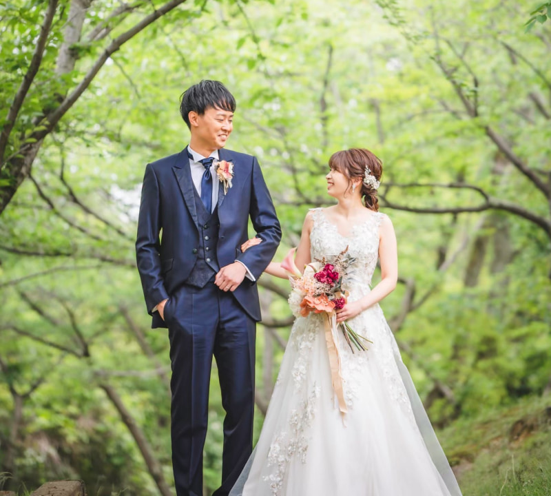 緑豊かな自然の中で、濃いネイビーブルーのスリーピーススーツを着た新郎と白いレースのウェディングドレスを着た花嫁が互いを見つめ合い、笑顔を交わしている様子。花嫁は華やかなブーケを手に持ち、背景には明るい緑の葉が茂る木々が映えている
