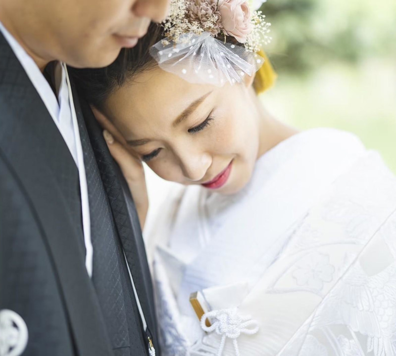 伝統的な和装の新郎新婦が寄り添う親密なポートレート。白い打掛を着た花嫁が黒い紋付羽織袴の新郎の肩に頭を預け、穏やかな笑顔を浮かべている