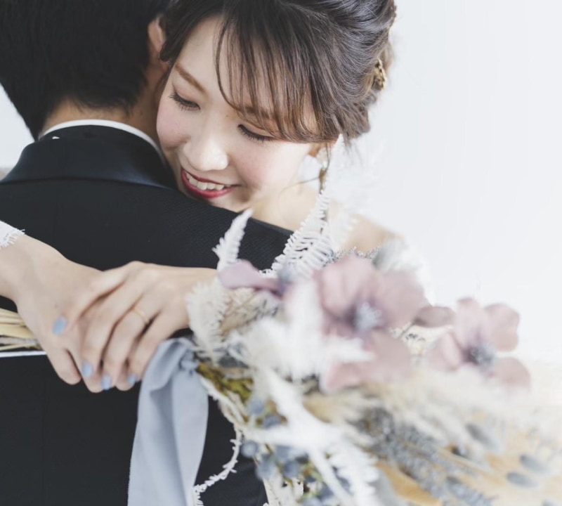 白いウェディングドレスを着た花嫁が、黒いタキシードを着た新郎を抱きしめている親密なクローズアップポートレート。花嫁は目を閉じ、満面の笑みを浮かべている