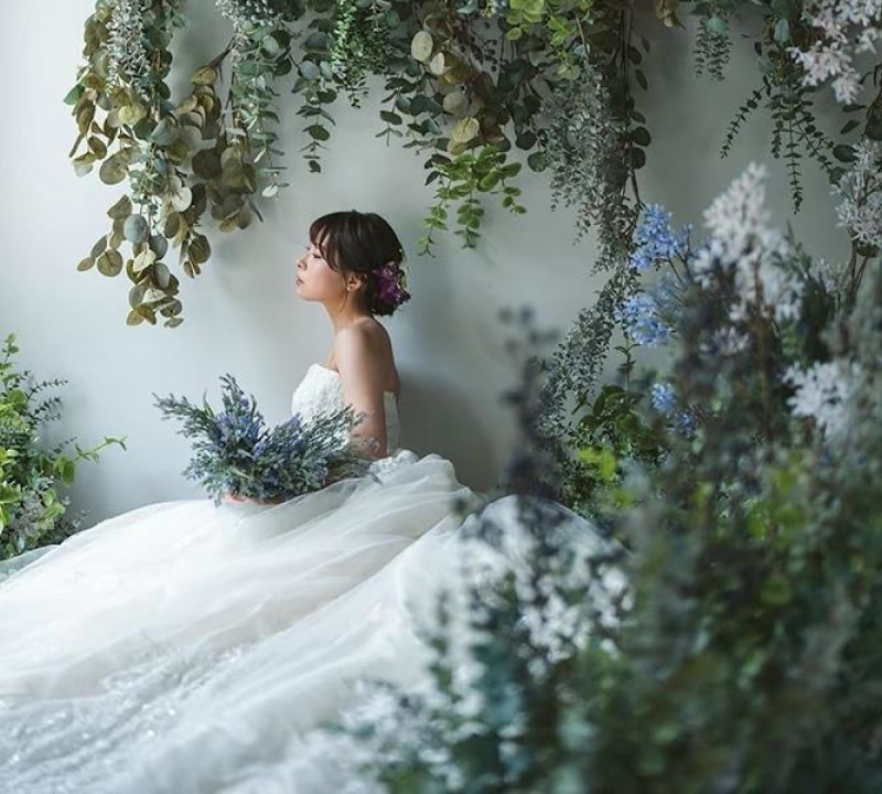 白いウェディングドレスを着た花嫁が、緑と白、淡い紫の花々で装飾された幻想的な空間に座り、手元のブーケを静かに見つめている横顔のポートレート