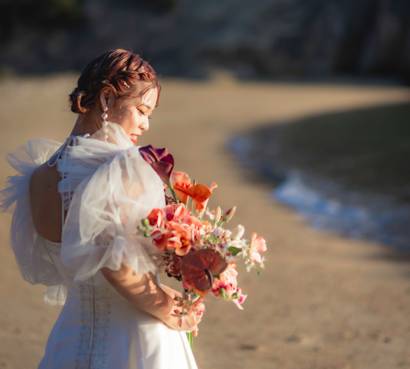 ウェディングドレスを着た女性が花束を持ち、静かなビーチでポーズをとっている。