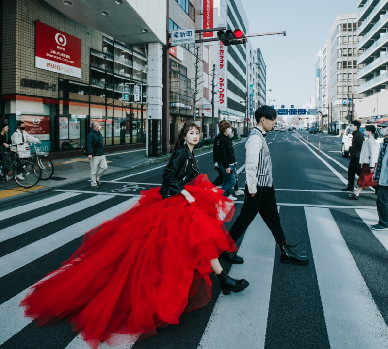 日本の都市の交差点で、鮮やかな深紅のチュールのロングドレスを着た女性と、白と黒のストライプ柄のベストを着た男性が横断歩道を渡っている様子。女性は黒いレザージャケットを羽織り、背景には「MUFG PLAZA」や「南新町」の看板が見える都市の風景が広がっている