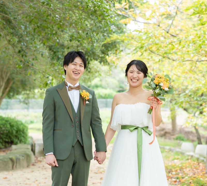 緑豊かな屋外で、オフホワイトのウェディングドレスを着た花嫁とオリーブグリーンのスリーピーススーツを着た新郎が手をつないで、カメラに向かって満面の笑みを浮かべている様子
