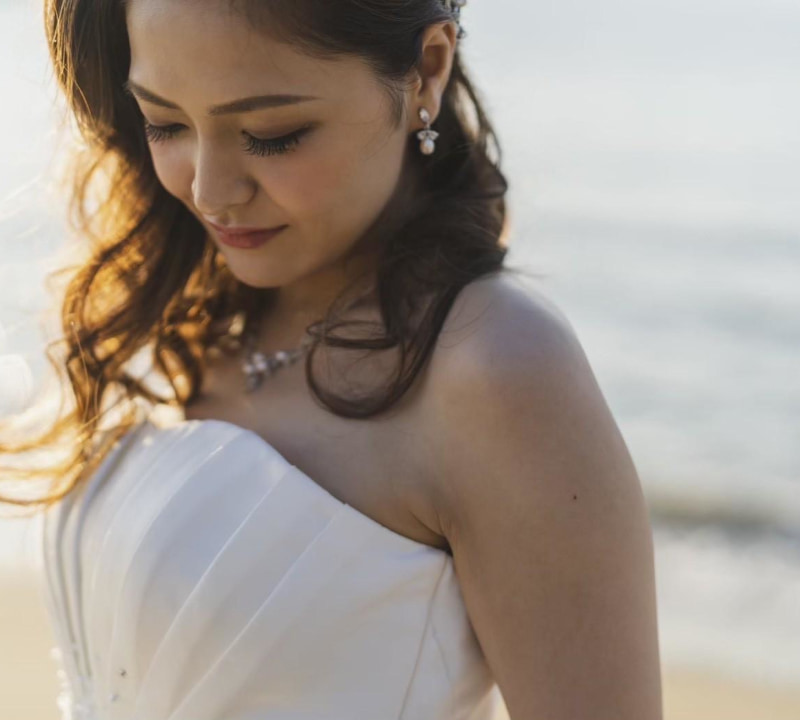 砂浜と海を背景に、白いウェディングドレスを着た花嫁のクローズアップ。花嫁は顔を伏せ、穏やかな笑顔を浮かべている