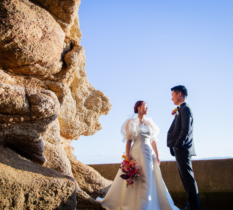 結婚式の衣装を着た新郎新婦が、岩の前で向かい合っているシーン。新婦は花束を持っている。