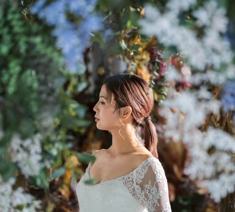 白いオフショルダーのウェディングドレスを着た花嫁の横顔のポートレート。髪はポニーテールにまとめられ、背景には緑豊かな花々がぼかして映っている