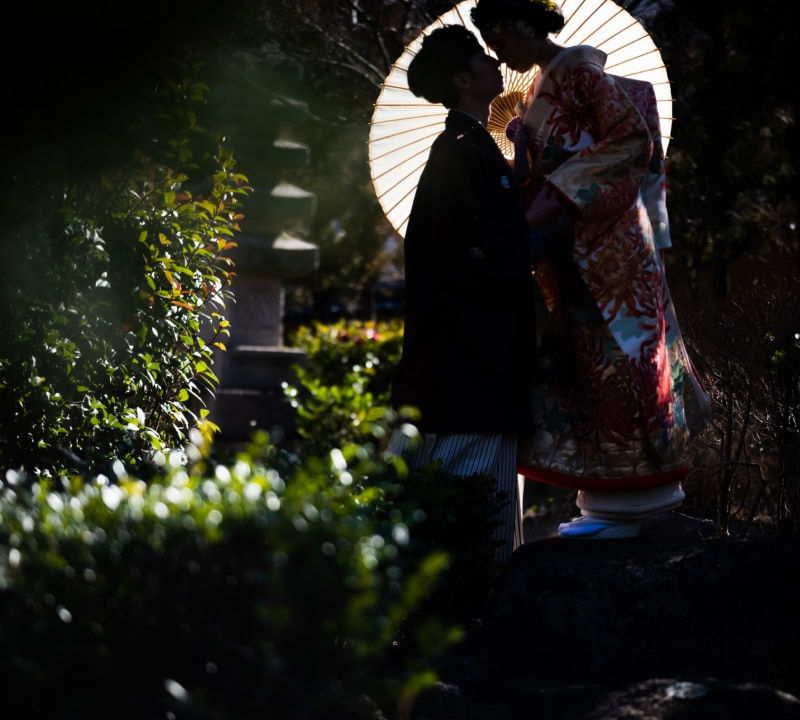 伝統的な和装の新郎新婦が、白い和傘を背景に顔を寄せ合い、互いを見つめ合っているロマンチックなシーン。新婦は華やかな色打掛を着用し、緑豊かな庭園で撮影されている