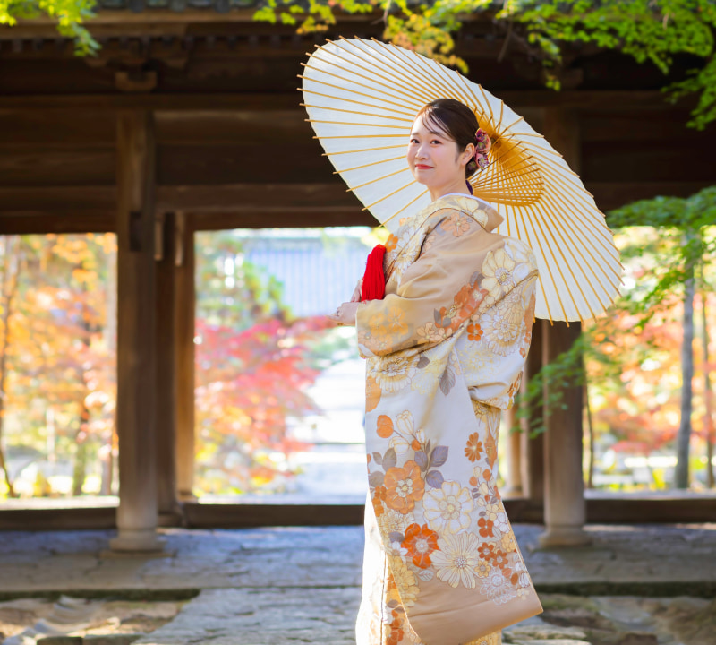 クリームやベージュ、金色の花模様が施された着物を着た若い女性が、白い和傘を右肩に乗せて立っている様子。髪には小さな花のヘアアクセサリーが飾られ、背景には暗い木製の門と鮮やかな紅葉が映えている