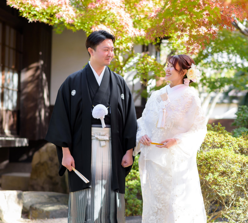 伝統的な和装を着た男性と女性が笑顔で向かい合っている。男性は黒い羽織と白い扇子を持ち、女性は白い着物に花飾りをつけている。