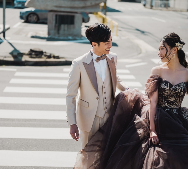 晴れた日の屋外で、横断歩道を歩きながら互いに顔を向け、笑顔を交わす新郎新婦。クリーム色のタキシードを着た新郎と、濃いブラウンまたはチャコールグレーのオフショルダーのウェディングドレスを着た花嫁が、都市の風景を背景に撮影されている