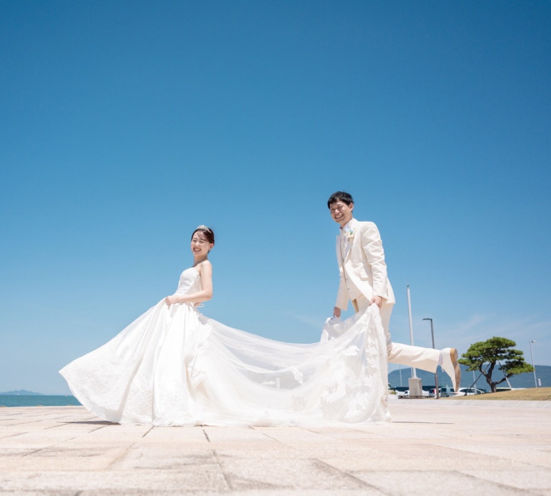 白いウェディングドレスを着た花嫁とクリーム色のタキシードを着た新郎が、青い海と空を背景に、笑顔でポーズをとっている結婚式の様子。新郎は花嫁のドレスの裾を持ち上げている