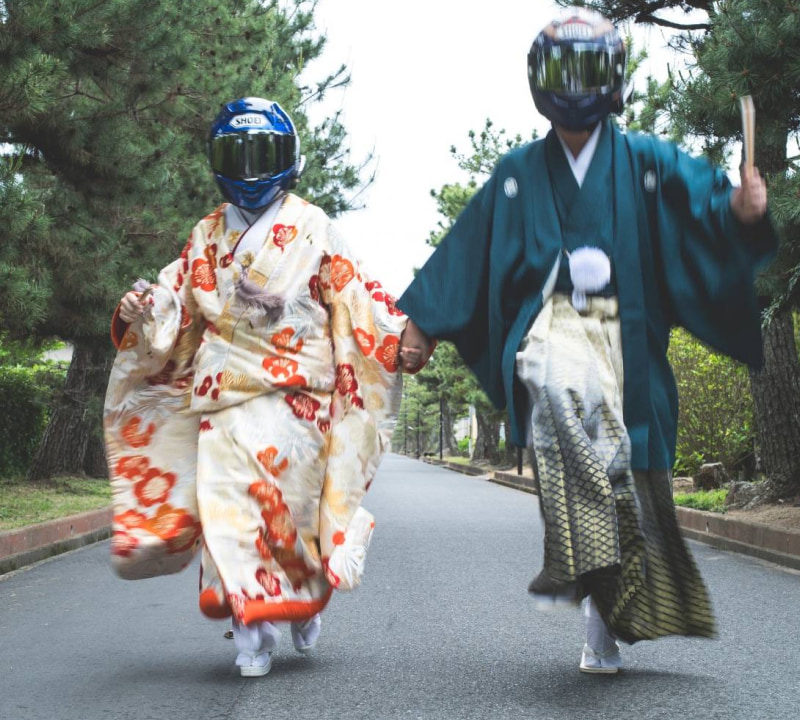 伝統的な着物を着た二人の人物が手をつないで走っている様子。ヘルメットを着用している。