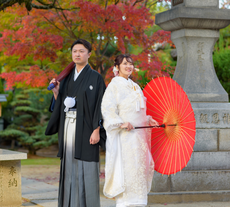 和装を着た男性と女性が赤い傘を持って立っている。背景には紅葉が見える。