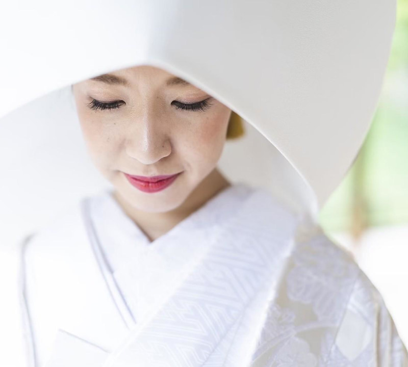 伝統的な白い打掛（白無垢）と大きな白い綿帽子を着用した花嫁のクローズアップポートレート。花嫁は目を伏せ、穏やかな微笑みを浮かべている