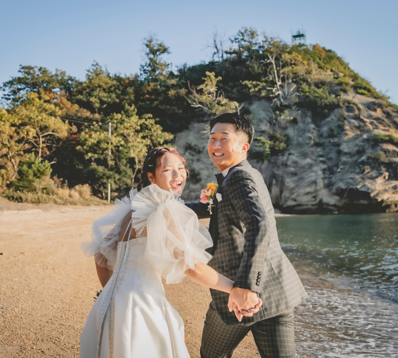 砂浜と海を背景に、白いウェディングドレスを着た花嫁とチェック柄のスーツを着た新郎が、緑豊かな木々と岩の崖の前で満面の笑みを浮かべている海岸線での結婚式の様子