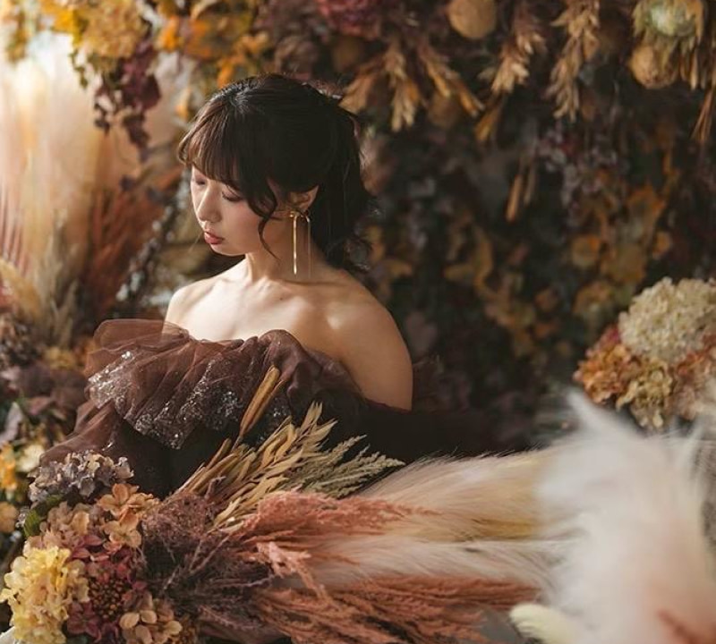 茶色のオフショルダードレスを着た女性が、ドライフラワーとパンパスグラスで飾られた秋らしい背景と前景の中で、うつむいて立っているポートレート