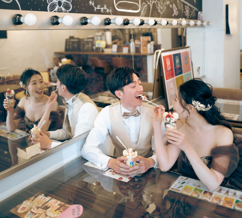 カフェで向かい合って座り、笑顔でデザートを楽しむ若いカップル。女性がスプーンでデザートをすくい、男性の口元に運んでいる瞬間を捉えた、明るく楽しい雰囲気のポートレート