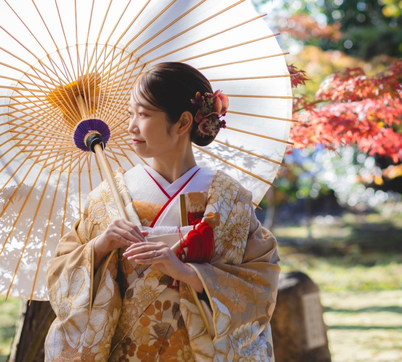 金色がかったベージュまたは薄茶色の着物を着た若い女性が、白い和傘を左肩に乗せて立っている様子。着物には大きな様式化された花（菊や牡丹など）の模様がクリーム、白、濃い金色/オレンジ色で施されている。髪は優雅なアップスタイルで、赤、オレンジ、紫の花のヘアピン（かんざし）が飾られている。白い和傘には竹の骨組みがはっきりと見え、紫色の装飾帯が付いている。背景には鮮やかな赤やオレンジの紅葉が映え、秋の庭園や神社の境内のような雰囲気が表現されている