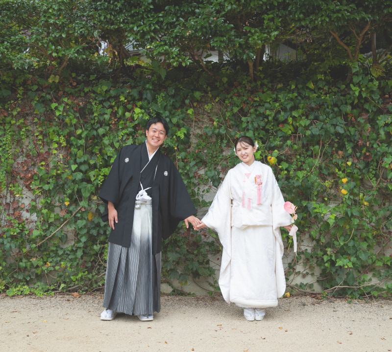 伝統的な和装を着た男女が手をつないで立っている。背景には緑の植物が生い茂っている。