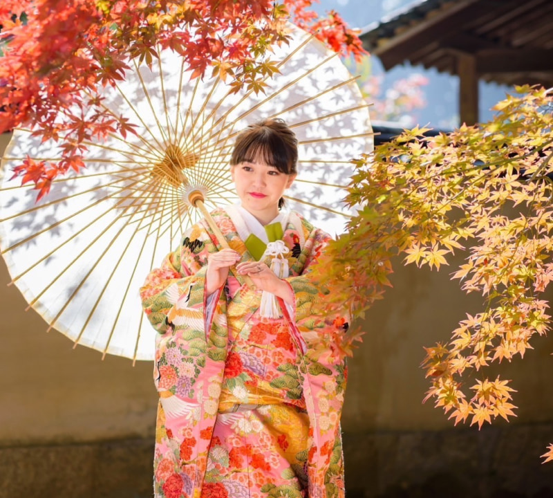 色とりどりの着物を着た女性が和傘を持ち、紅葉の中に立っている。