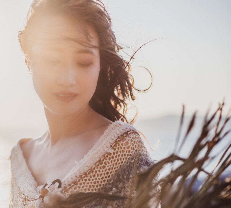 強い逆光の中で、目を閉じて風になびく髪を持つ若い女性のクローズアップポートレート。クリーム色のニットのようなトップスを着用し、木製のような留め具と羽毛のような装飾が胸元に見える