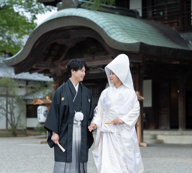伝統的な日本の木造建築を背景に、和装の結婚衣装をまとった新郎新婦が手を取り合い、互いに見つめ合って笑顔を交わしている様子。花嫁は白無垢を着用している
