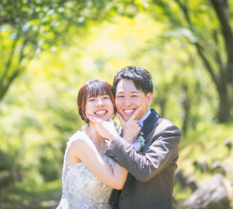 結婚式の衣装を着たカップルが笑顔でポーズをとっている写真。背景には緑の木々が広がっている。