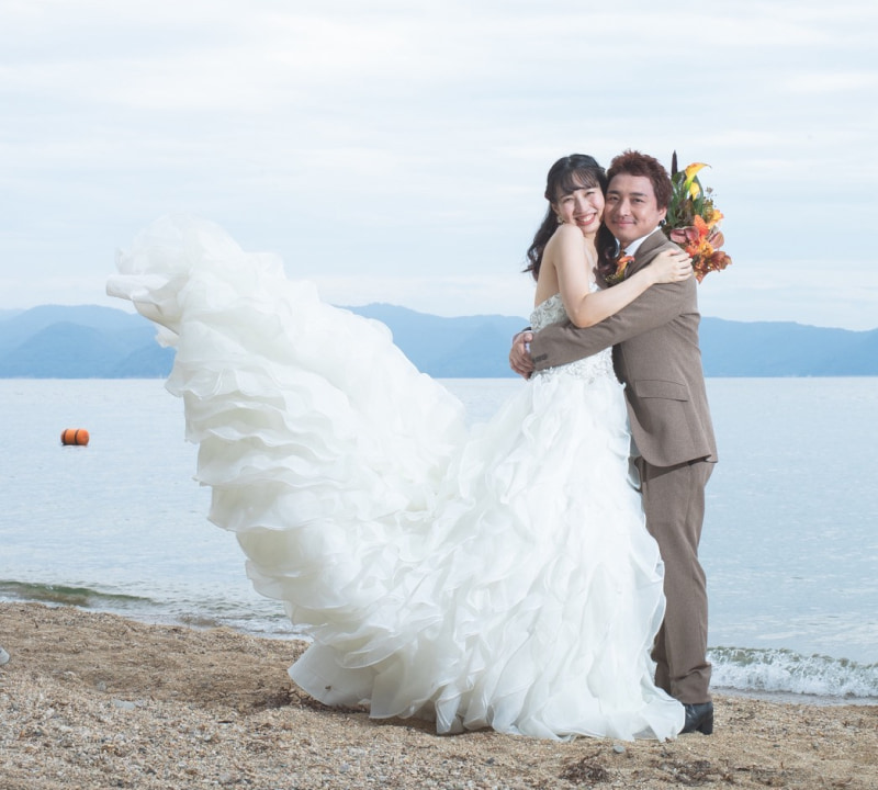 海辺でウェディングドレスを着た新婦とスーツを着た新郎が抱き合っている。ドレスはフリルが特徴的で、背景には穏やかな海が広がっている。